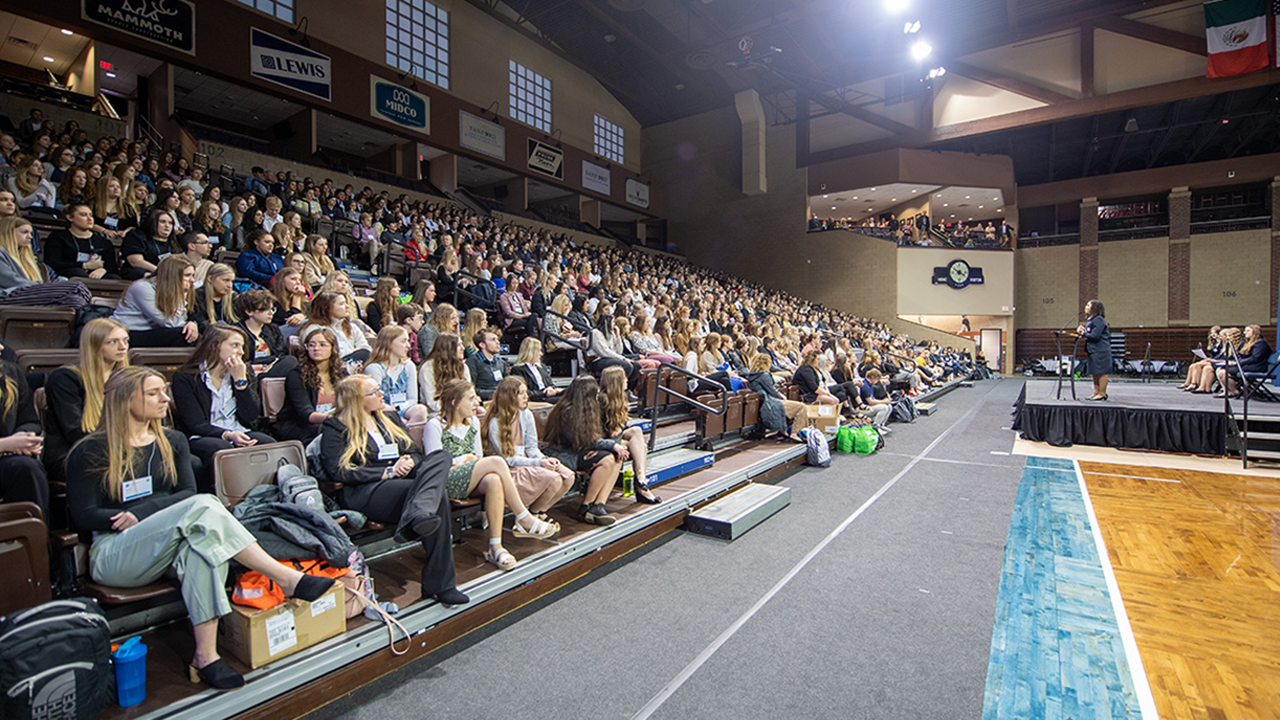 Hundreds of students sit in bleachers in a gym and listen to a speaker.