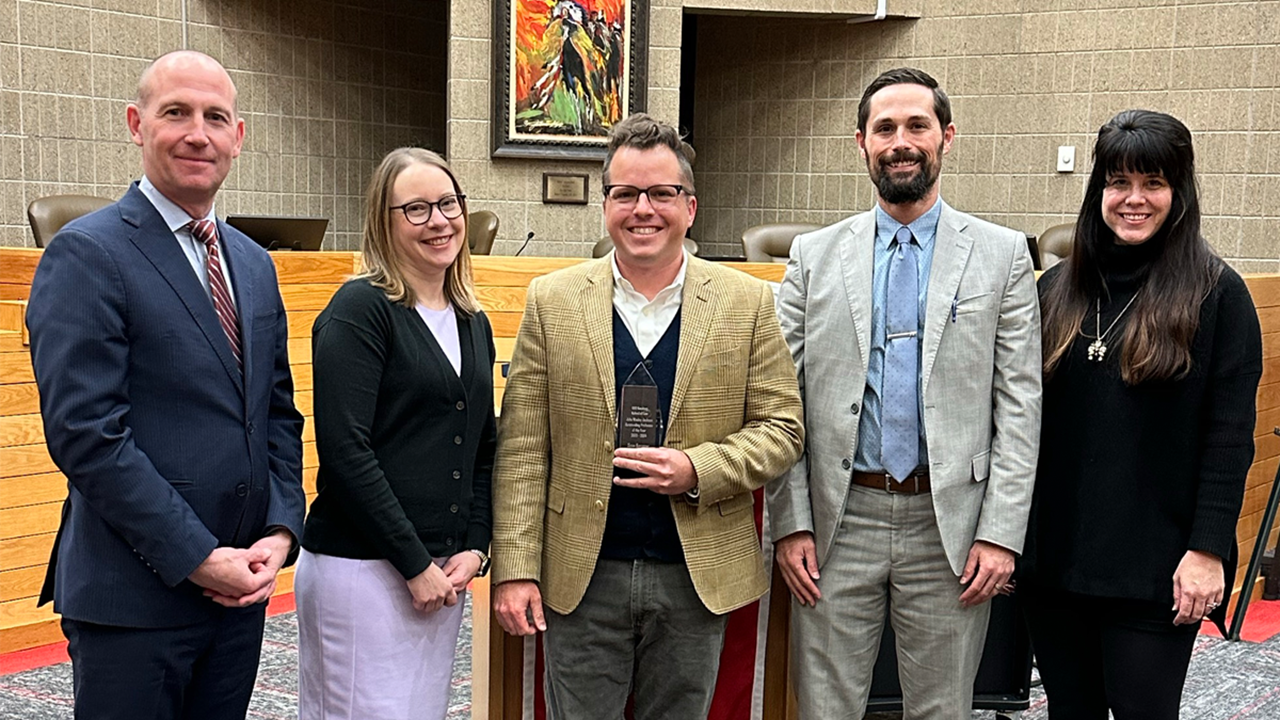 Sean Kammer holds his award and smiles for a photo in the law school courtroom. There are two people standing to his left and two to his right.