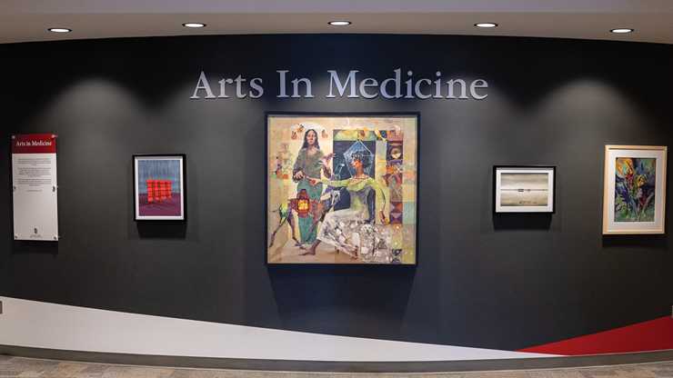 A curved wall that has five framed artwork pieces hanging on it. Above the artwork is the words Arts In Medicine.