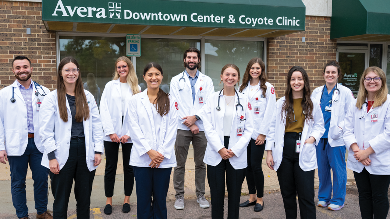 10 medical students wearing white coats stand together in front of a building. The building has a green awning with the text Avera Downtown Center & Coyote Clinic printed on it.