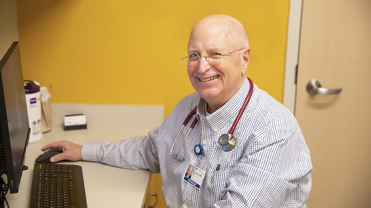 Dr. Tom Hermann has a stethoscope around his neck and sits at a desk in a doctors office.