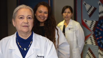 Dr. Sophie Two Hawk wears a white coat and stands in front of Dr. Arna Mora and Dr. Carol Whitman.