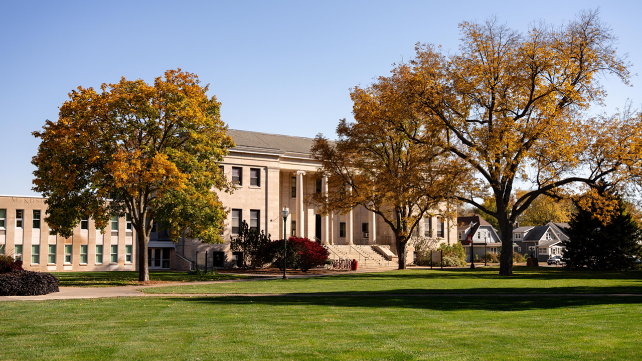 The University of South Dakota’s College of Arts & Sciences building framed by trees with golden autumn leaves, with a bright blue sky overhead.