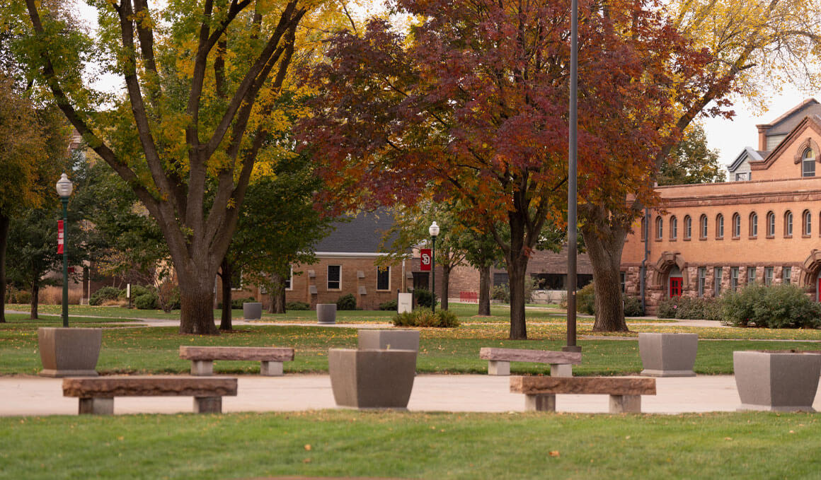 A peaceful campus scene in autumn featuring large trees with yellow, orange and red leaves, stone benches arranged along a walkway, and historic brick buildings in the background.