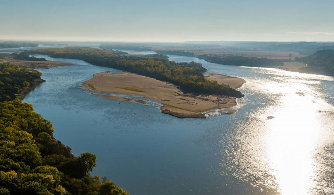 An aerial view of the Missouri River winding through a landscape of trees and sandbars under a clear blue sky, with sunlight reflecting off the water’s surface.