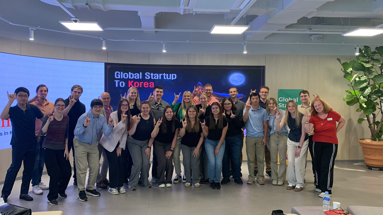 Students from the Beacom School of Business pose for a group photo, with the presentation "Global Startup to Korea" in the background.