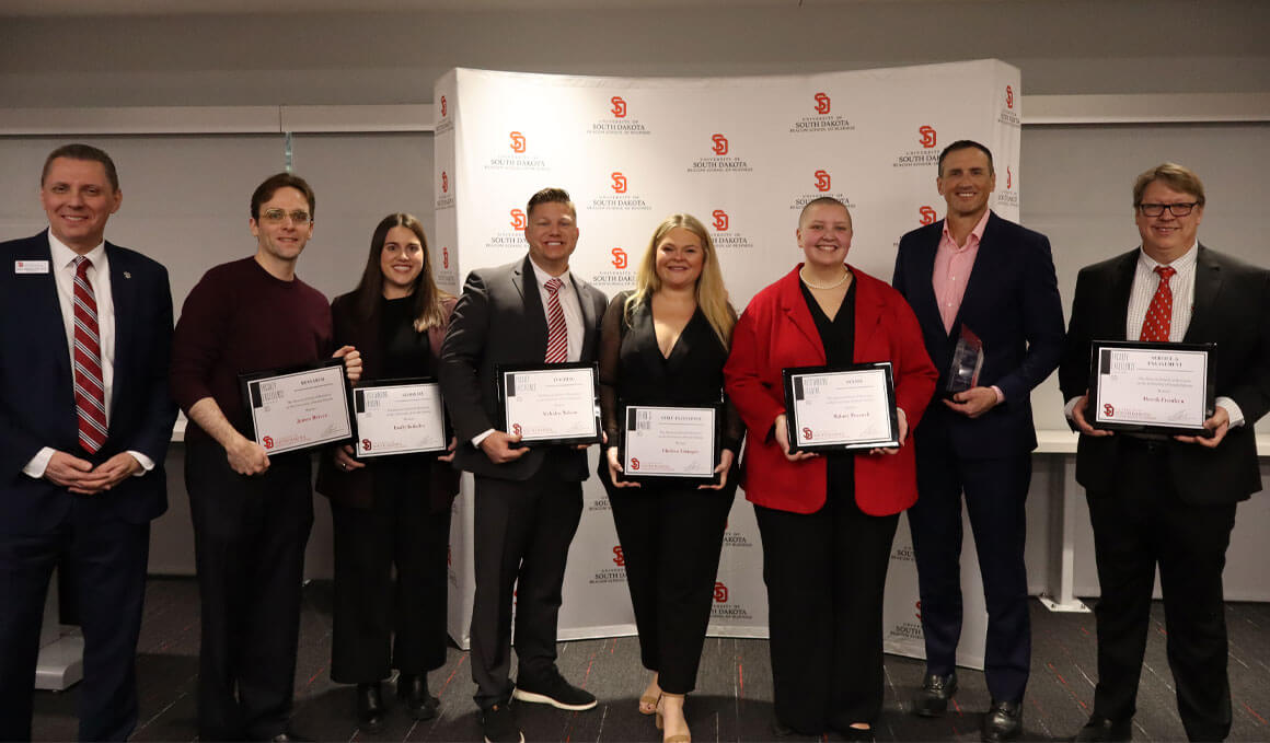 Staff, faculty and students from the Beacom School of Business stand together, holding their awards and smiling for the camera. They are standing in front of a gray and red Beacom School of Business backdrop, that contains the USD logo.