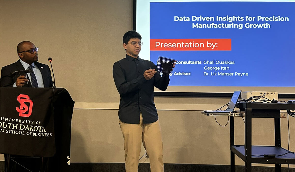 Two presenters lead a business presentation while one holds a demonstration item in front of a slide titled “Data Driven Insights for Precision Manufacturing Growth.”