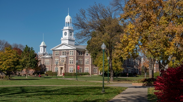 Old Main stands in the background of the USD campus, with fall colored trees and bushes amongst the landscaping.