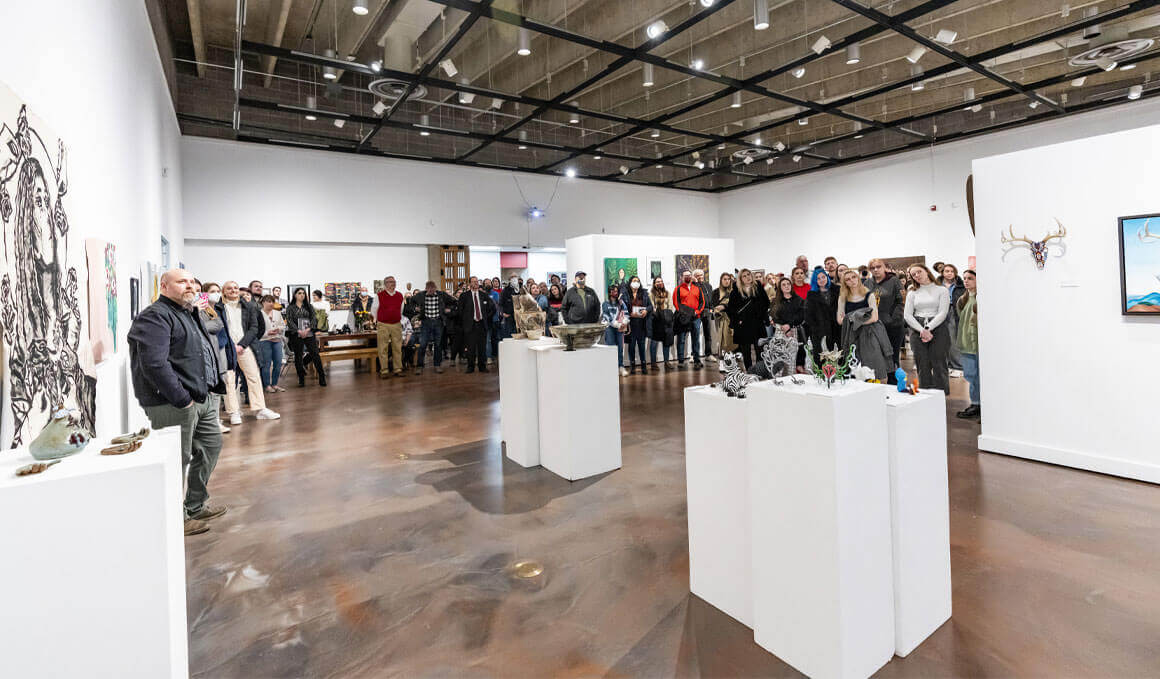 A crowd gathers at the USD Art Galleries during the Stilwell Student Awards Exhibition. The crowd is surrounded by artwork, placed on white walls and white tables, that's made from students.