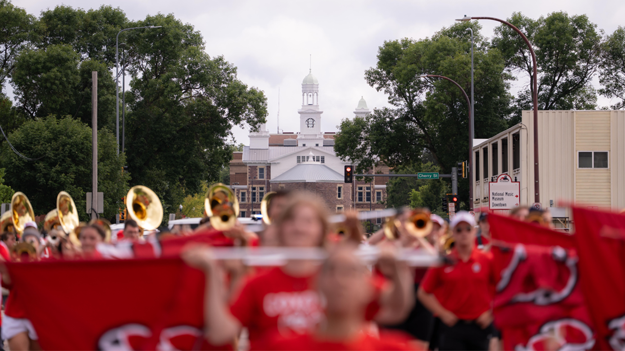 The SOUND of USD Marching Band marches in a parade wearing USD apparel and holding instruments, with baton twirlers in the forefront and Old Main in the background against white and gray sky.