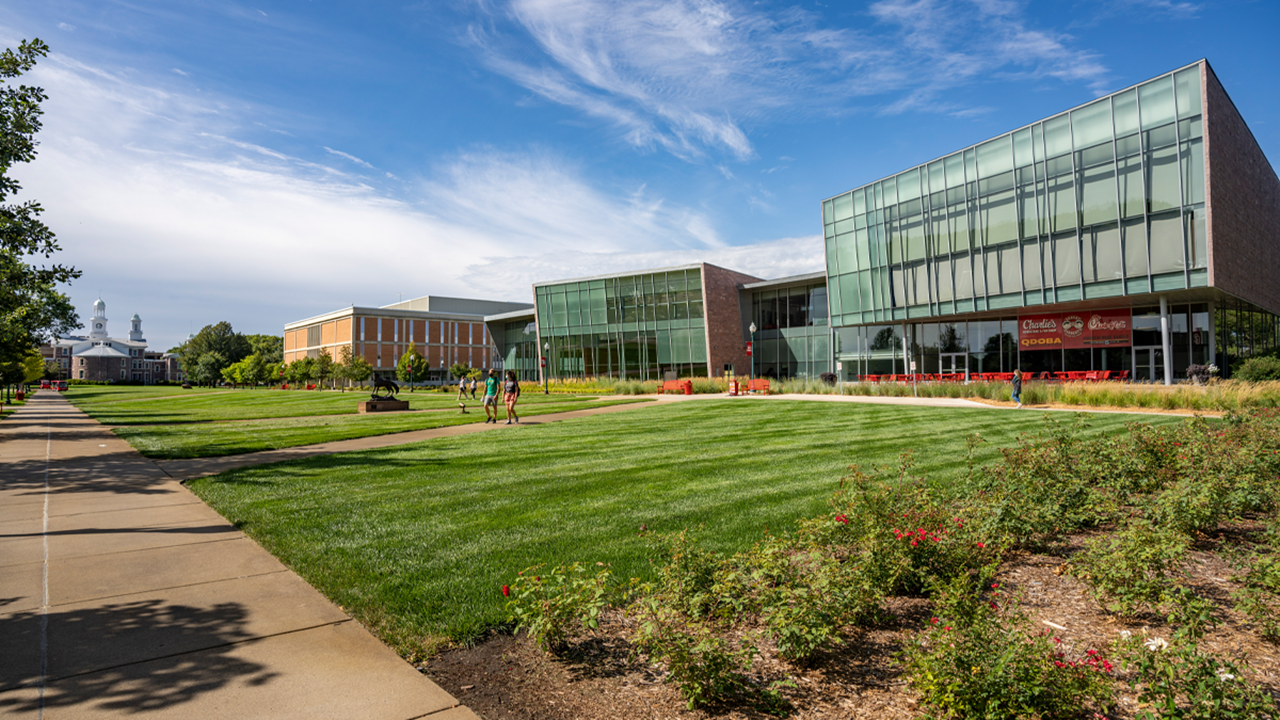 Students walk on the USD campus in front of the Muenster University Center, under a blue sky and next to bright green grass.