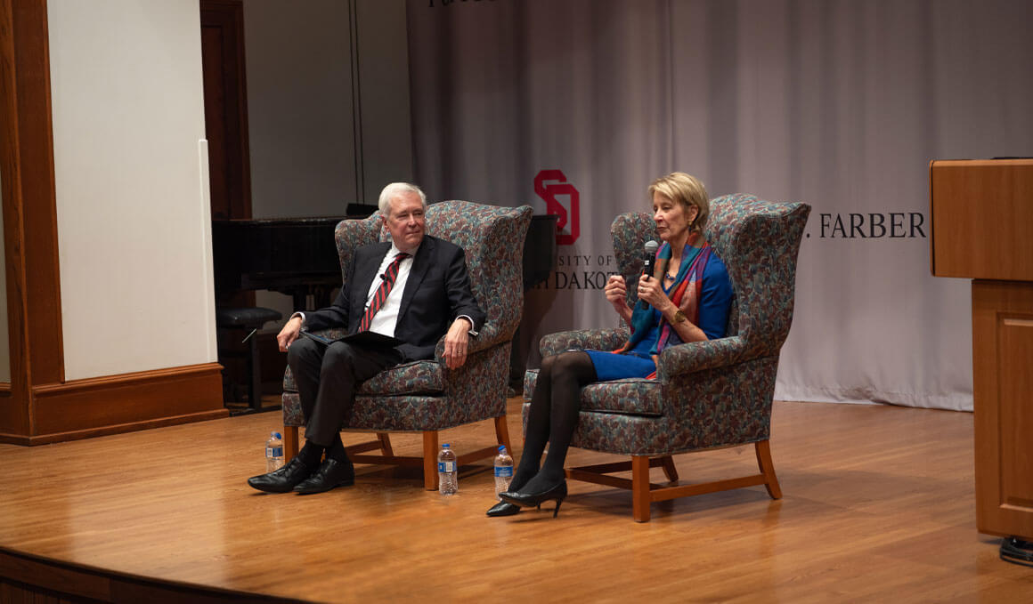 James and Deborah Fallows sit in big, oversized chairs on a stage, with James looking to his left at Deborah, who is holidng a microphone in her left hand and speaking to the audience.
