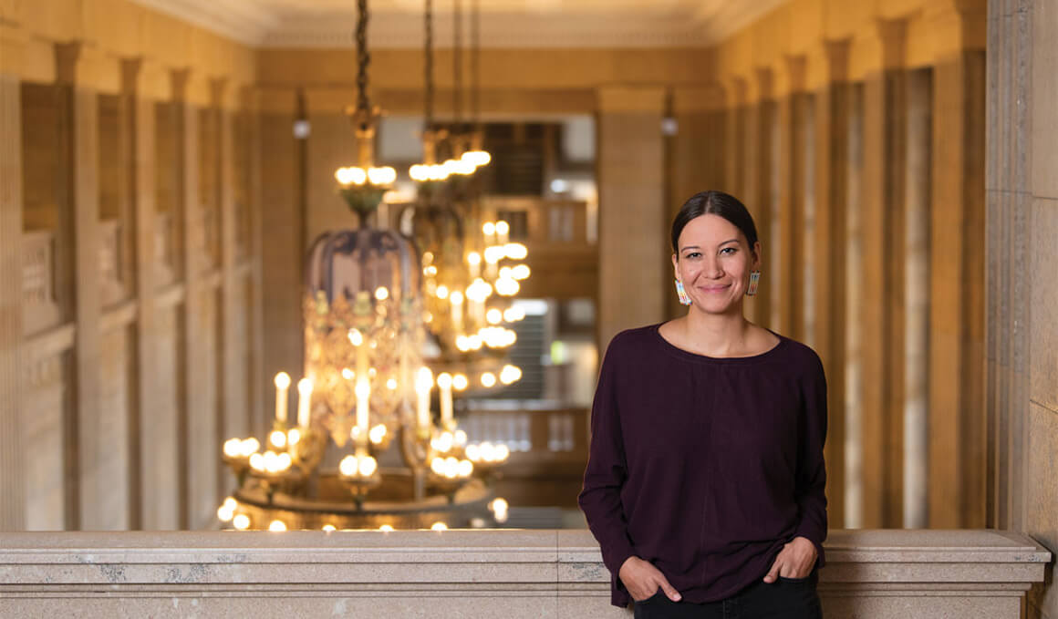 Megan Red Shirt-Shaw stands smiling in an elegant hallway with large chandeliers and warm lighting.