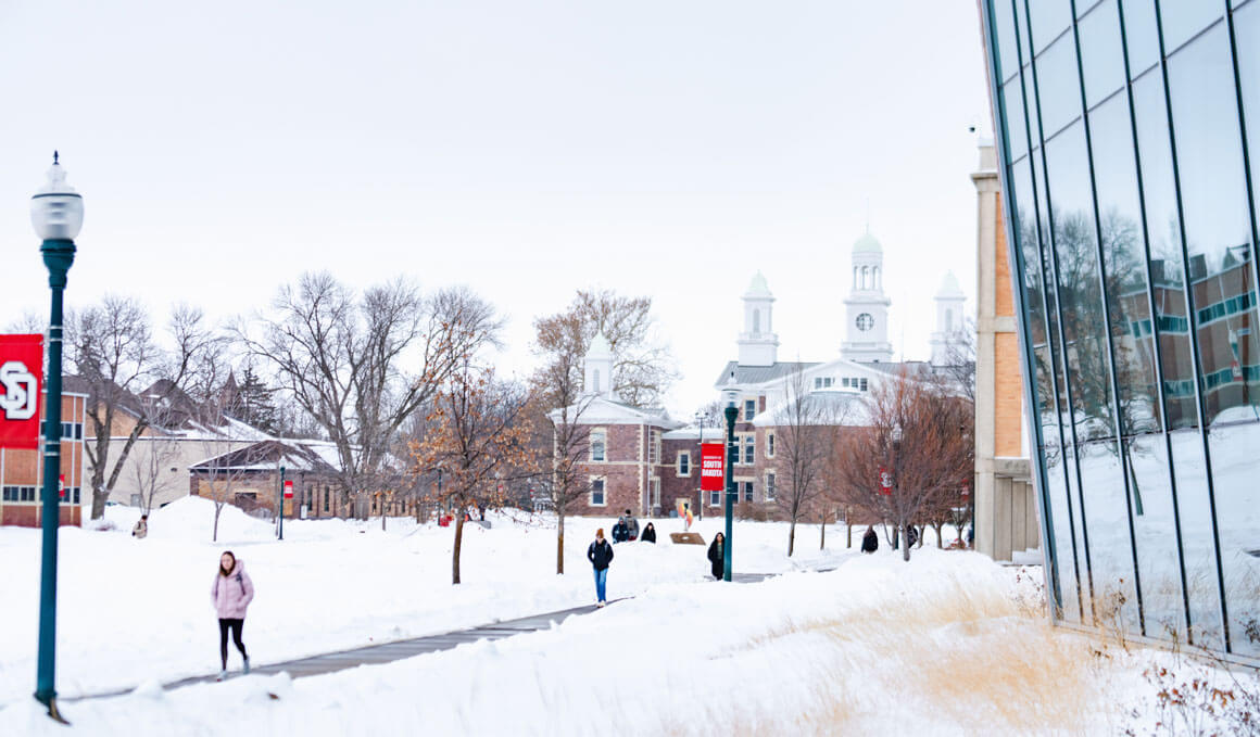 A snowy winter scene on the USD campus shows students walking along a path lined with bare trees, historic brick buildings with white cupolas and red USD banners.