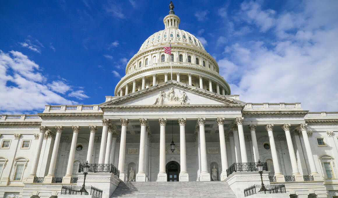 Exterior view of the U.S. Capitol Building in Washington, D.C., showcasing the white dome, columns and steps against a bright blue sky with white clouds.
