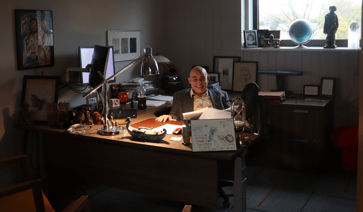 Michael McKey sits at a cluttered wooden desk in a dimly lit office, with a window and a variety of pictures and objects visible.