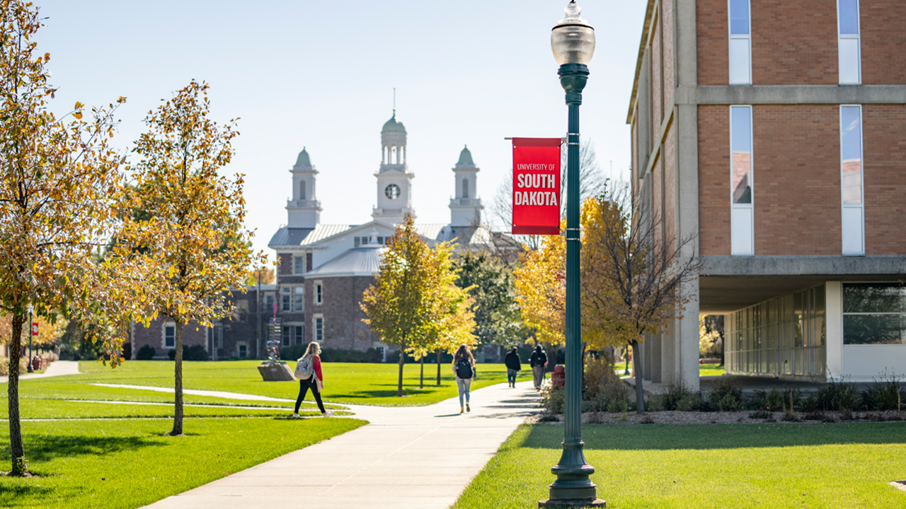 Students walk along a tree-lined sidewalk on the University of South Dakota campus, with Old Main visible in the background and a red USD banner on a lamppost in the foreground.