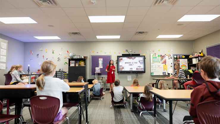 A USD student teacher stands in front of a SMART board and speaks to students in a classroom setting.