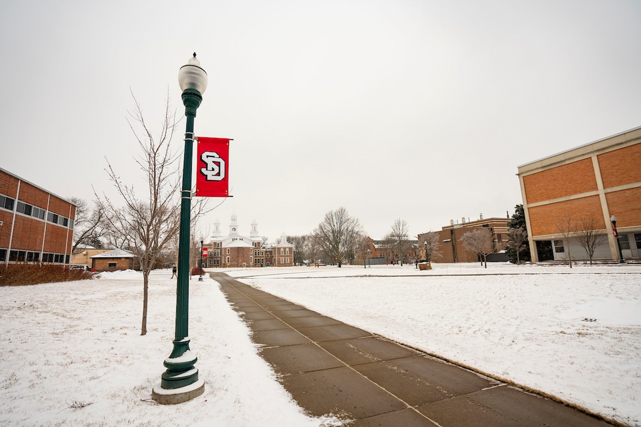 USD school banner attached to a light pole near sidewalk on a snowy campus.