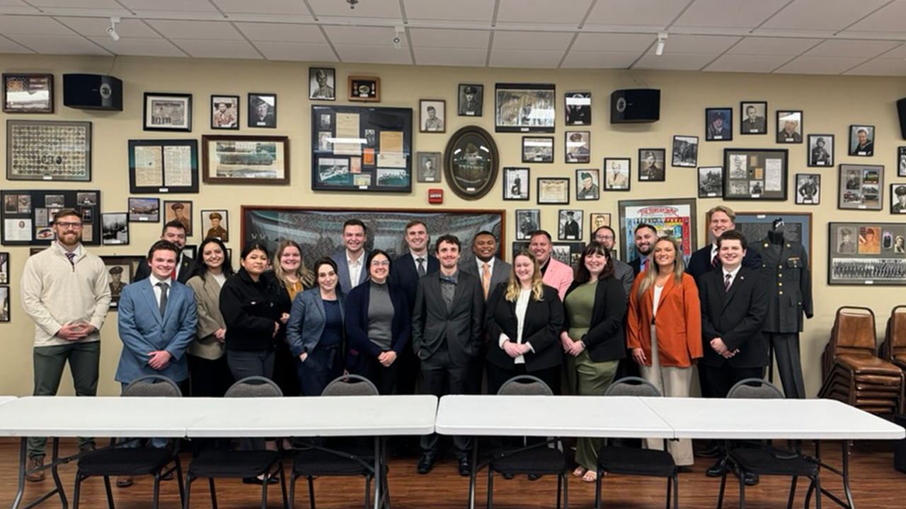USD Knudson School of Law students gather for a group photo, in a conference room at the Veteran Legal Education Group, in front of a wall that has a myriad of photos on it.