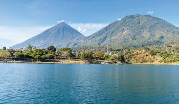 Guatemalan landscape with ocean water in the forefront, and mountains, trees and buildings in the background.