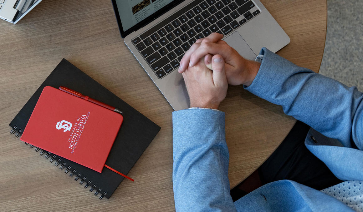 A student sits at a table with his laptop and notebook, one of which has the USD Beacom School of Business logo on it.