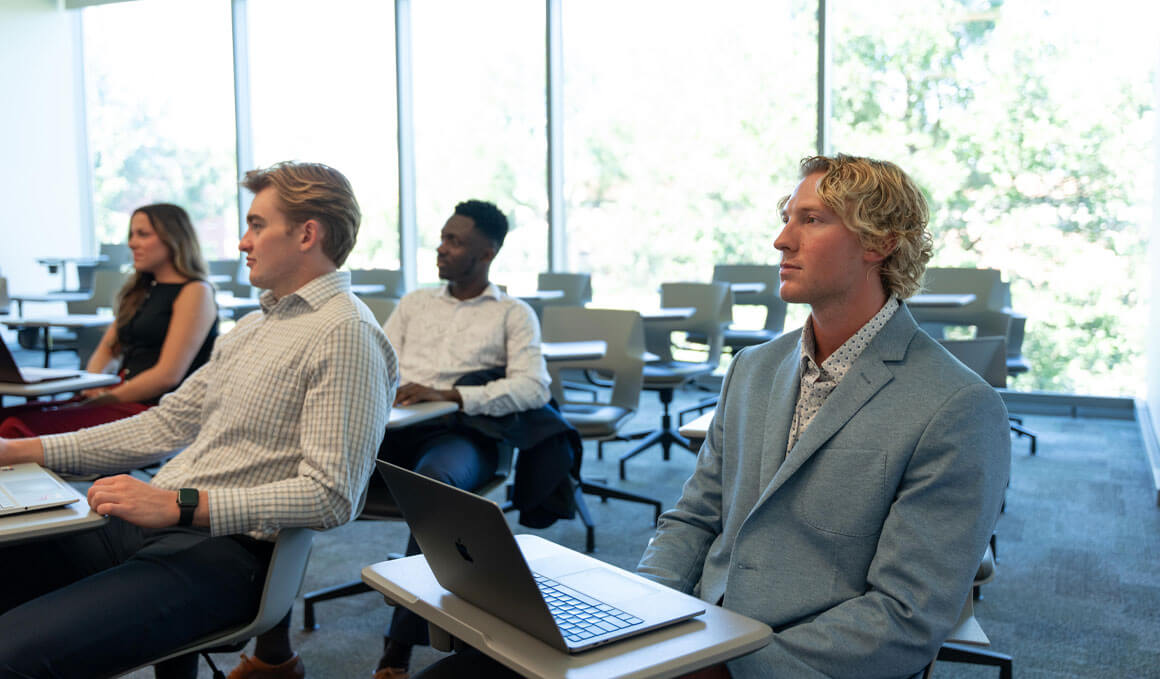 USD Beacom School of Business students sit in desks, in a classroom setting. They are looking off in the distance, towards an instructor.
