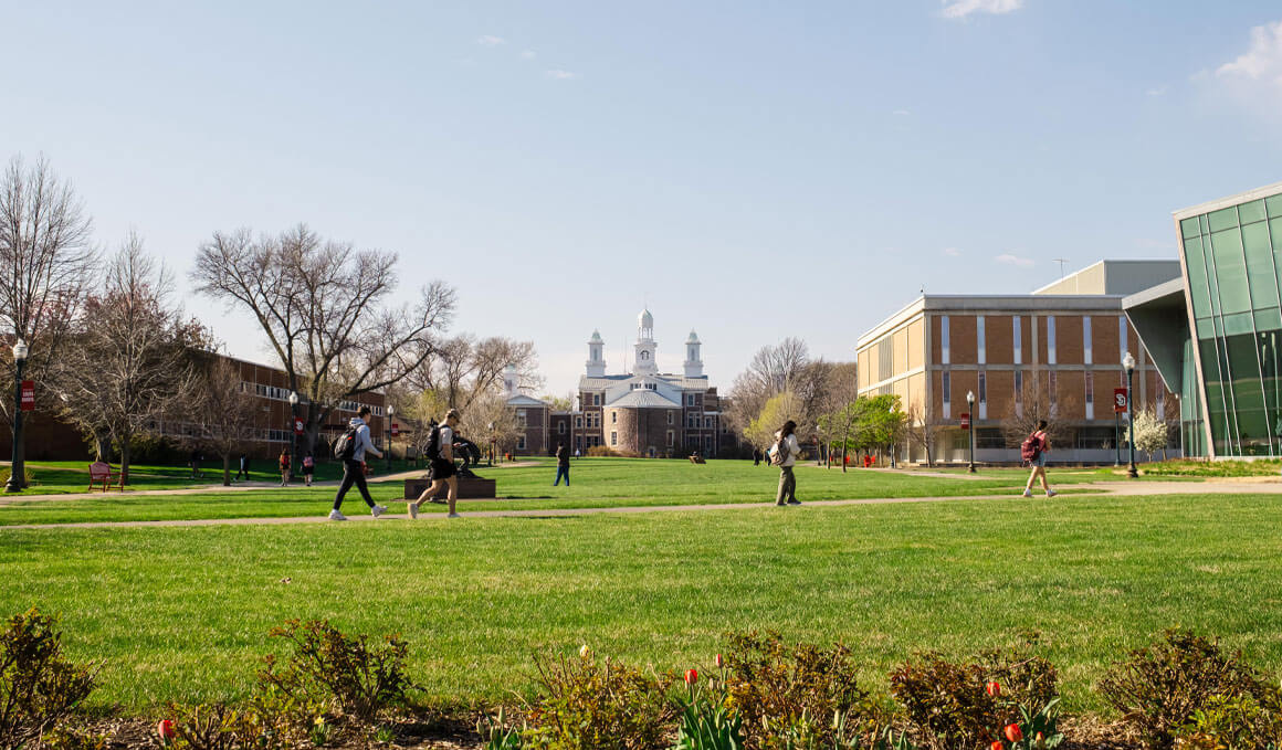 This photo shows a center view of the USD campus, looking towards Old Main, with the Muenster University Center to the right of it. There are many students walking on the sidewalks.