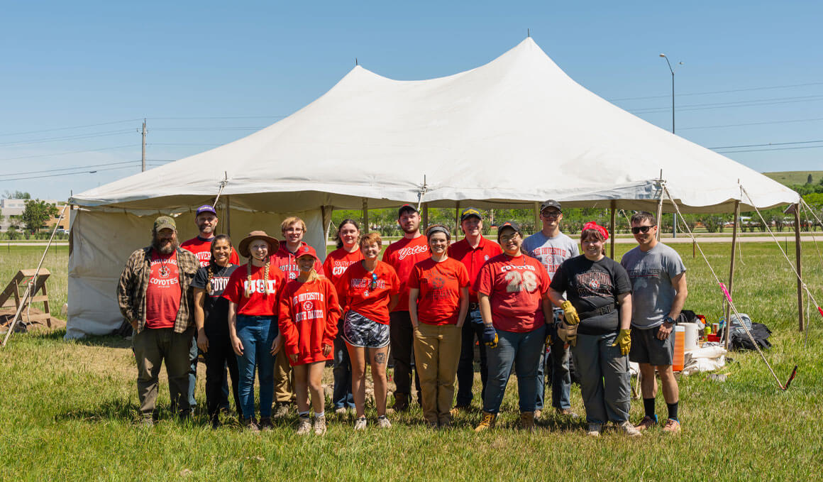 The 2026 USD Archaeology Field School stands in a remote, grassland area. They are dressed in USD apparel and archaeology attire, standing in front of a white tent, smiling at the camera.