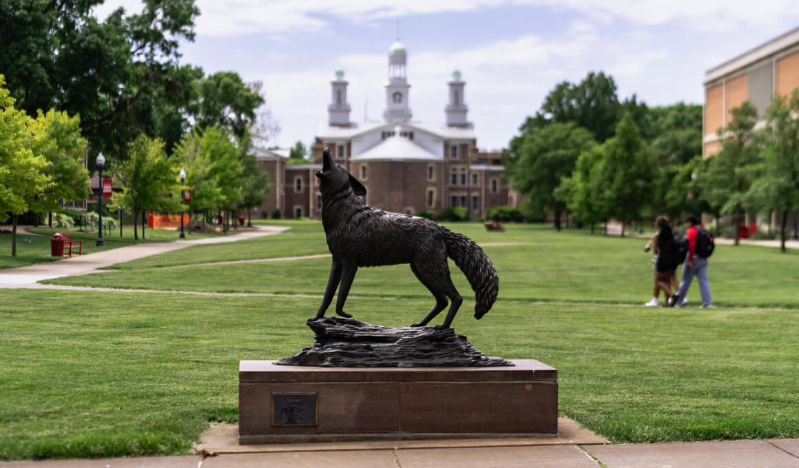 This image shows a close-up of the Coyote Legacy statue, which sits in the middle of the USD campus. Students are in the background, walking through the campus and Old Main can be seen.