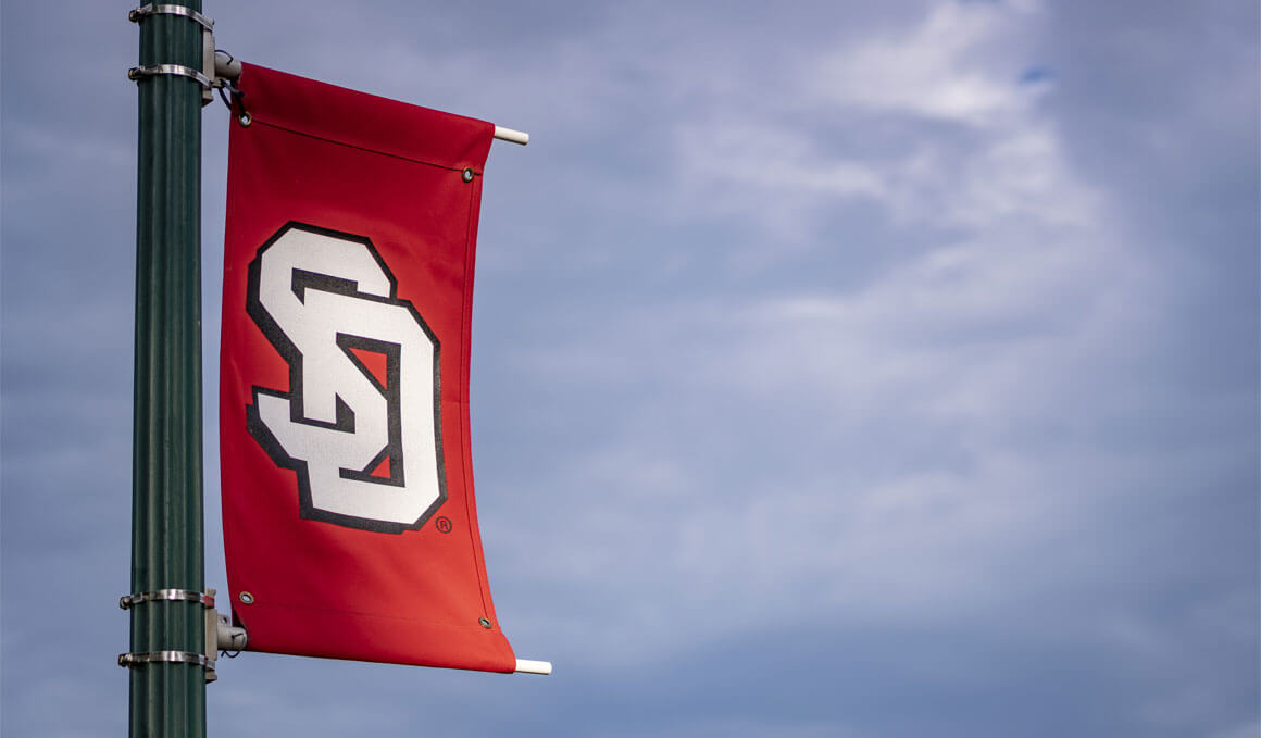 A red University of South Dakota banner, with the white letters "SD" clings to a light pole with a blue, semi-cloudy sky in the background.