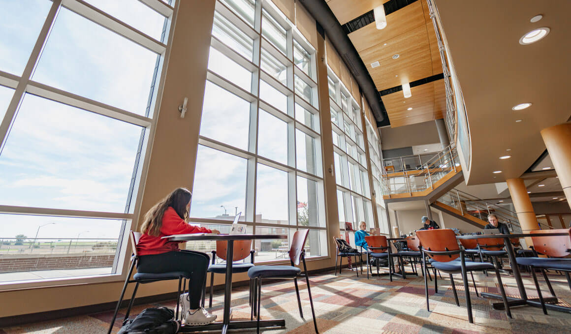 A USD – Sioux Falls student sits in a commons area at a table studying. Behind the student are floor to ceiling windows, showing the rest of the USD – Sioux Falls campus.
