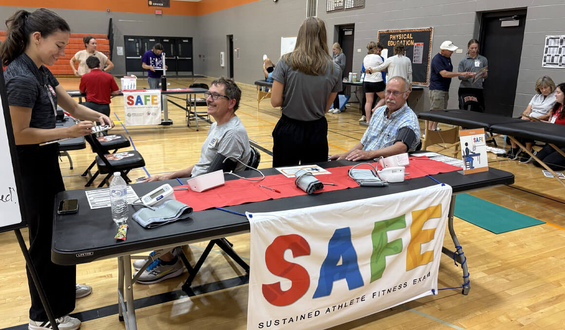 This photo shows a view of a fitness health check station in a gym, showing participants seated at a table and having their blood pressure measured.