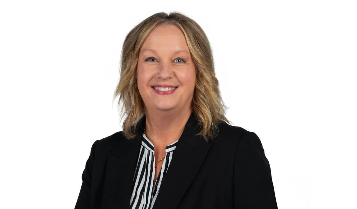 Nicky Gilbertson sits for her professional photo. She is wearing a black blazer with a striped shirt and smiling at the camera.