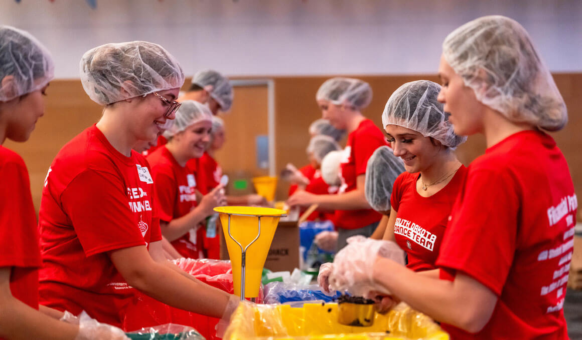 USD students stand in a line as they participate in Feed the Funnel. They are wearing hair nets and bright red USD and/or Coyote gear, as well as shirts that say "Feed the Funnel."