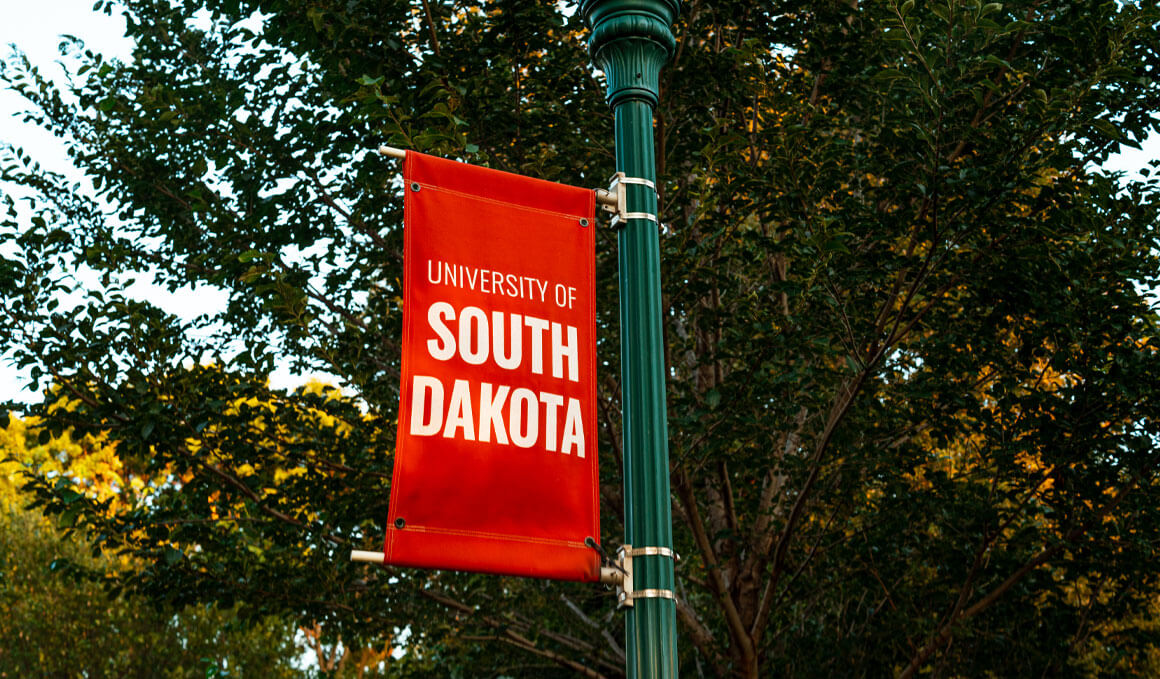 A close-up of a red and white banner that reads, "University of South Dakota." It is attached to green lamp post with trees in the background.
