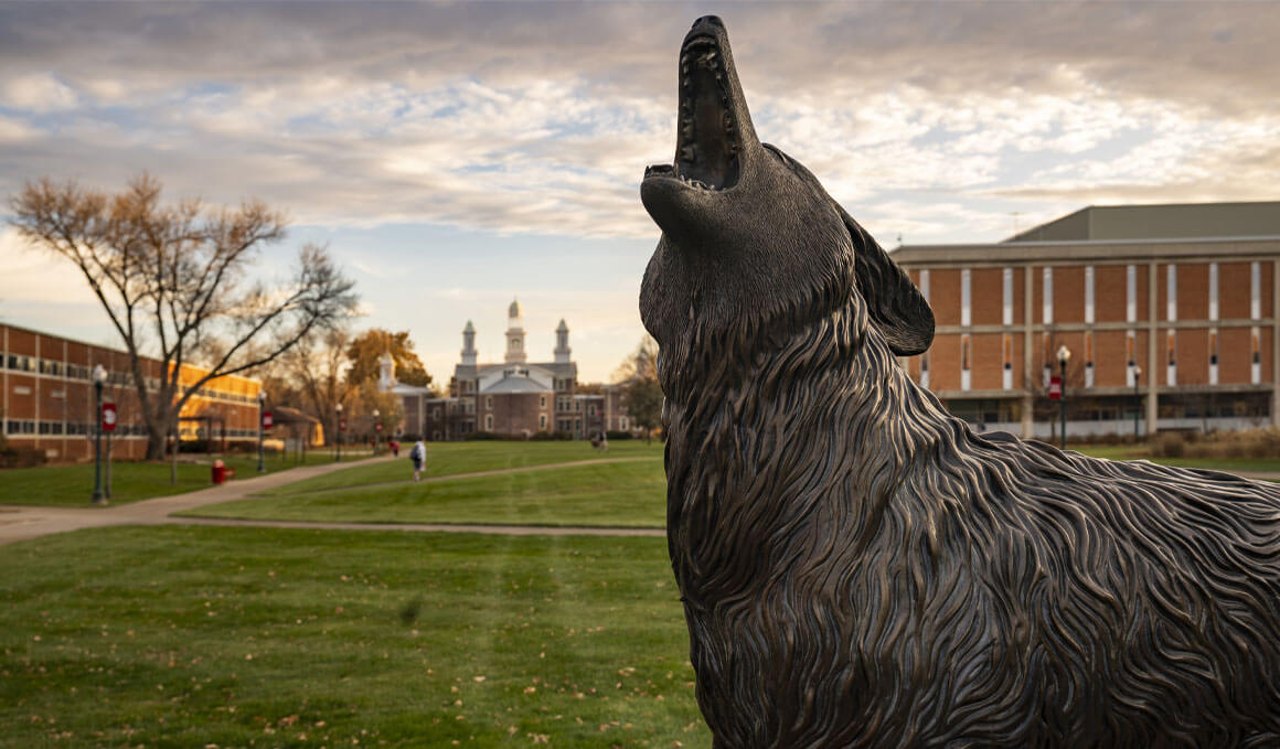 The Coyote Legacy Statue stands on the USD campus lawn, with brick campus buildings and a cloudy sunset sky behind it.