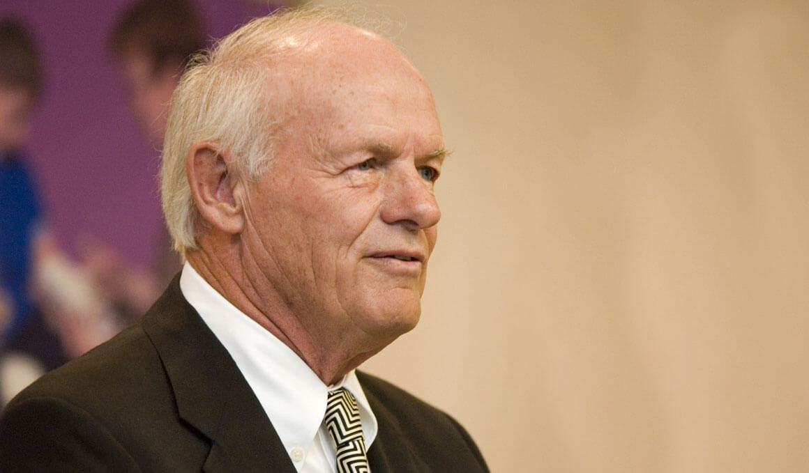 T. Denny Sanford stands at an event, smiling and looking off into the distance. He is standing in front of what appears to be a beige wall, with a purple poster on it.