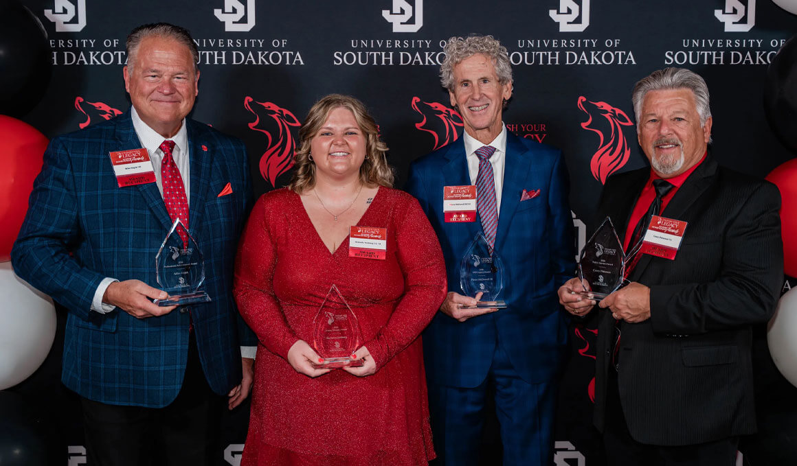 Four USD alumni pose with awards before a University of South Dakota backdrop, smiling in formal attire at the Unleash Your Legacy Awards event ceremony.