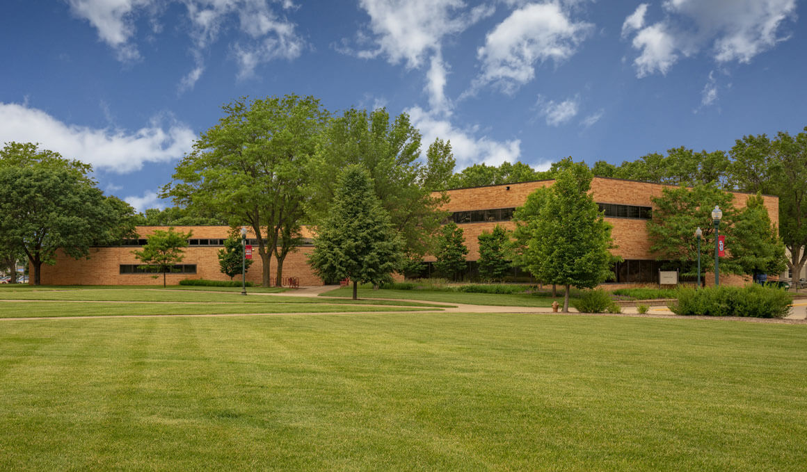 The Knudson School of Law sits on the University of South Dakota campus. Surrounding it are blue skies with a few clouds, green grass and trees, along with lightpoles that have a USD banner attached to them.