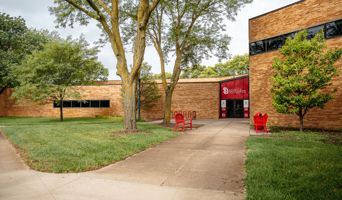 The Knudson School of Law building sits on the USD campus, surrounded by grass, trees, a concrete walkway and bright red benches by the entrance.