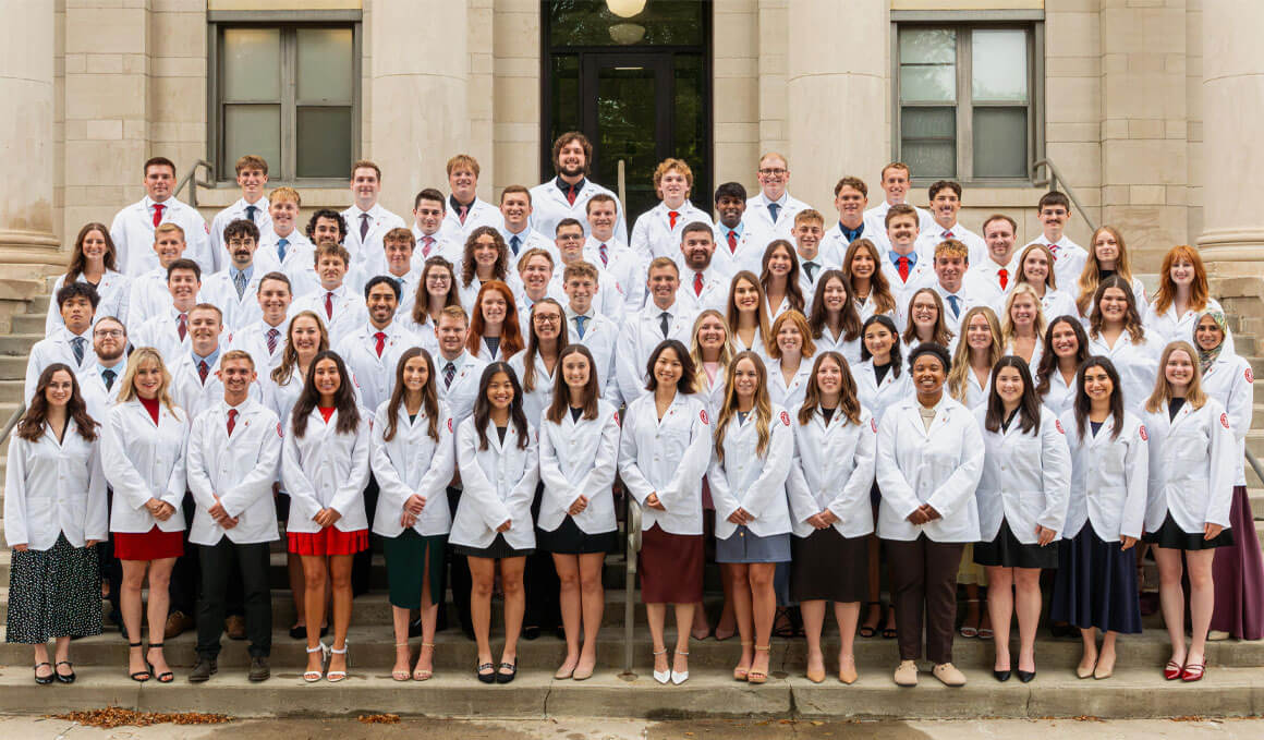 The Sanford School of Medicine's Class of 2030 stands on front steps of a USD building in their white coats.