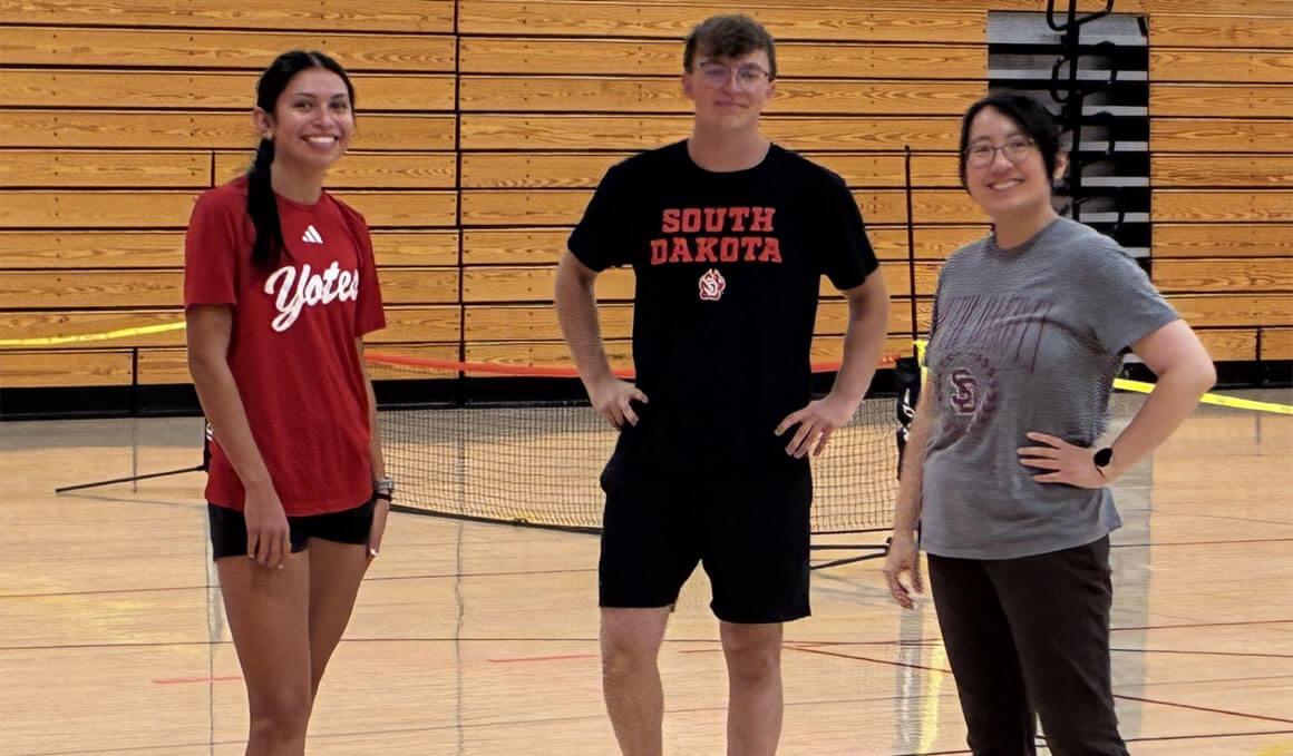 Three USD KSM students stand in a school gym. Behind them are nets and bleachers. They are wearing USD attire and smiling at the camera.