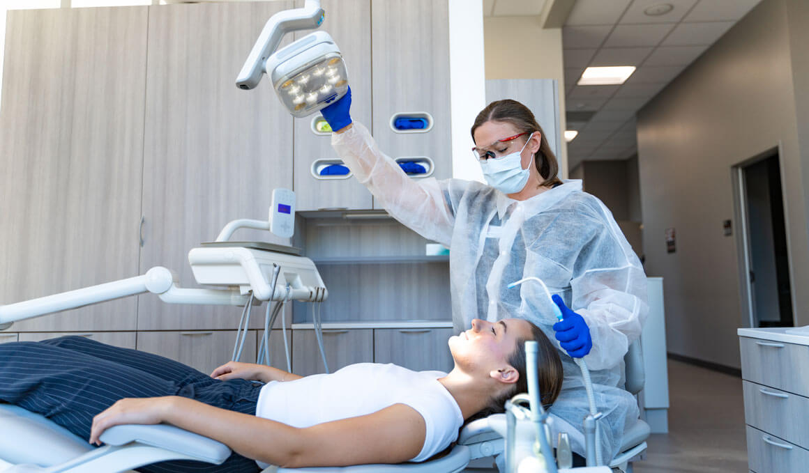 A USD student stands in a mock-dental setting, wearing full protective gear and adjusting the light on an overhead exam light while treating a patient during a clinical session.