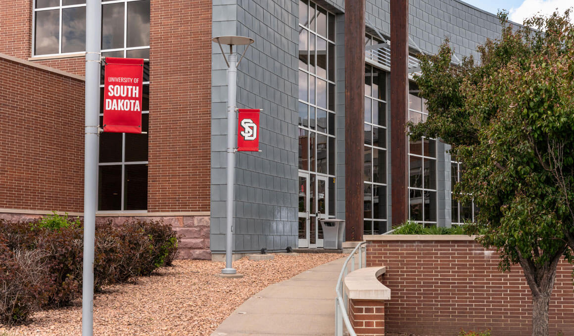 Exterior of a University of South Dakota – Sioux Falls building with brick and gray stone walls, large windows, landscaping, and red USD banners.