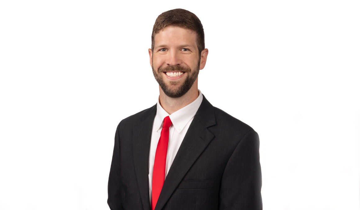Jed Droge sits for his professional photo against a white backdrop. He is wearing a black suit and red tie and smiling at the camera.
