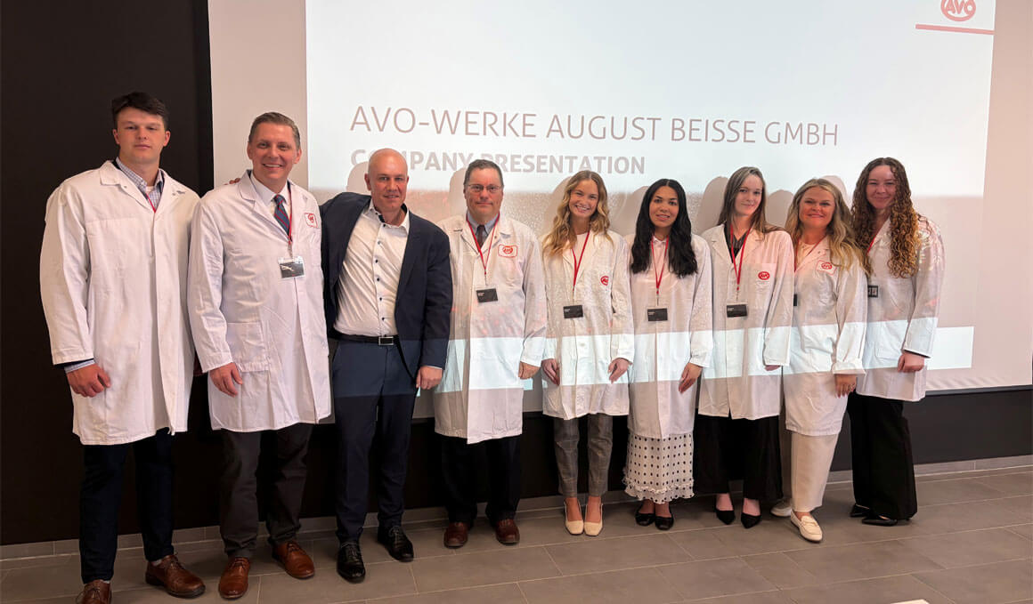 Members of the trade mission trip stand in front of a screen, wearing long white lab coats. They are smiling at the camera.