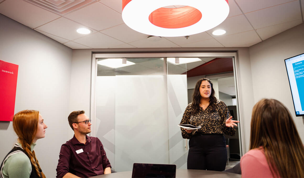 A USD student stands and gestures while leading a professional discussion with three other students seated around a circular table in a modern office space.