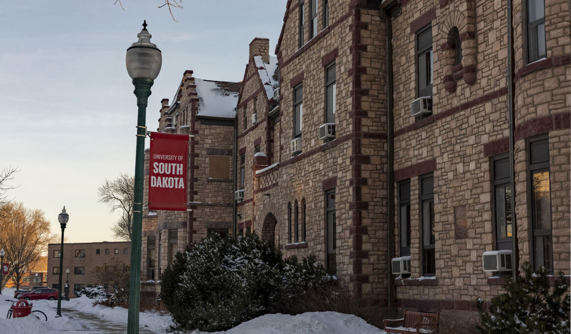 A side profile of East Hall on the USD campus, with snow on the ground and a lightpole in the foreground that is holding a red banner that says, "University of South Dakota."